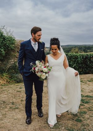 A couple walks on a hilltop path during their wedding day, with the bride wearing white and groom in navy suit.