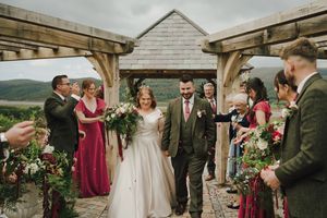 Bride and groom walk down the aisle at an outdoor rustic wooden pergola wedding ceremony with guests holding floral arrangements.