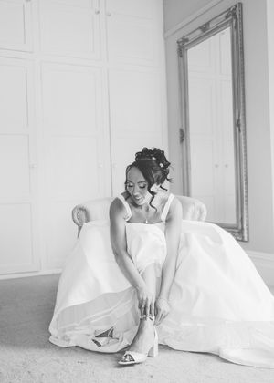 Black and white photo of a bride in a white gown sitting in a chair adjusting her heels near an ornate mirror.