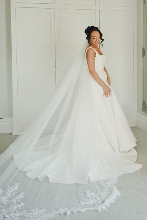 Bride in elegant white ballgown with long lace-trimmed cathedral veil, posing in bright white room.