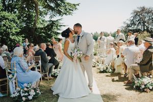 Bride and groom share first kiss at outdoor wedding ceremony with seated guests and floral decorations in sunny garden.
