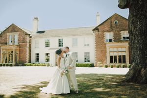 Bride and groom sharing a kiss outside an elegant brick manor house on their wedding day.