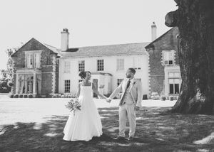 Black and white photo of a bride and groom holding hands outside an elegant country house wedding venue.
