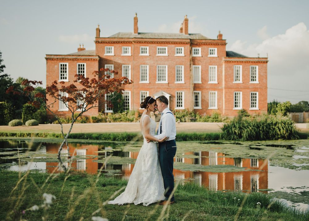 Bride and groom embrace before a grand red brick manor house reflected in a pond on their wedding day.