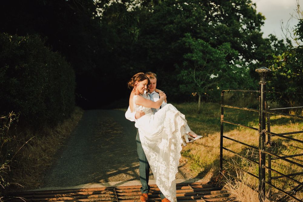 Groom carrying bride in white lace dress on a sunlit country path surrounded by lush green trees.