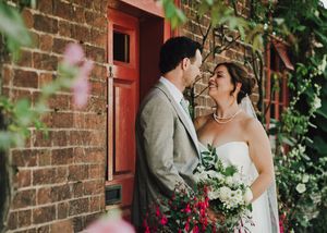 Bride and groom share a romantic moment by a red door on a brick wall, surrounded by lush greenery and flowers.