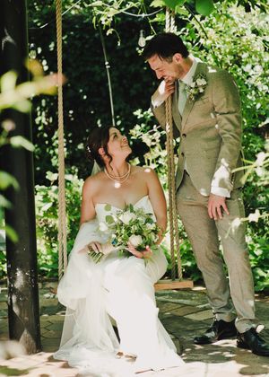 Bride sitting on a garden swing holding a bouquet, smiling up at groom in tan suit surrounded by lush greenery.