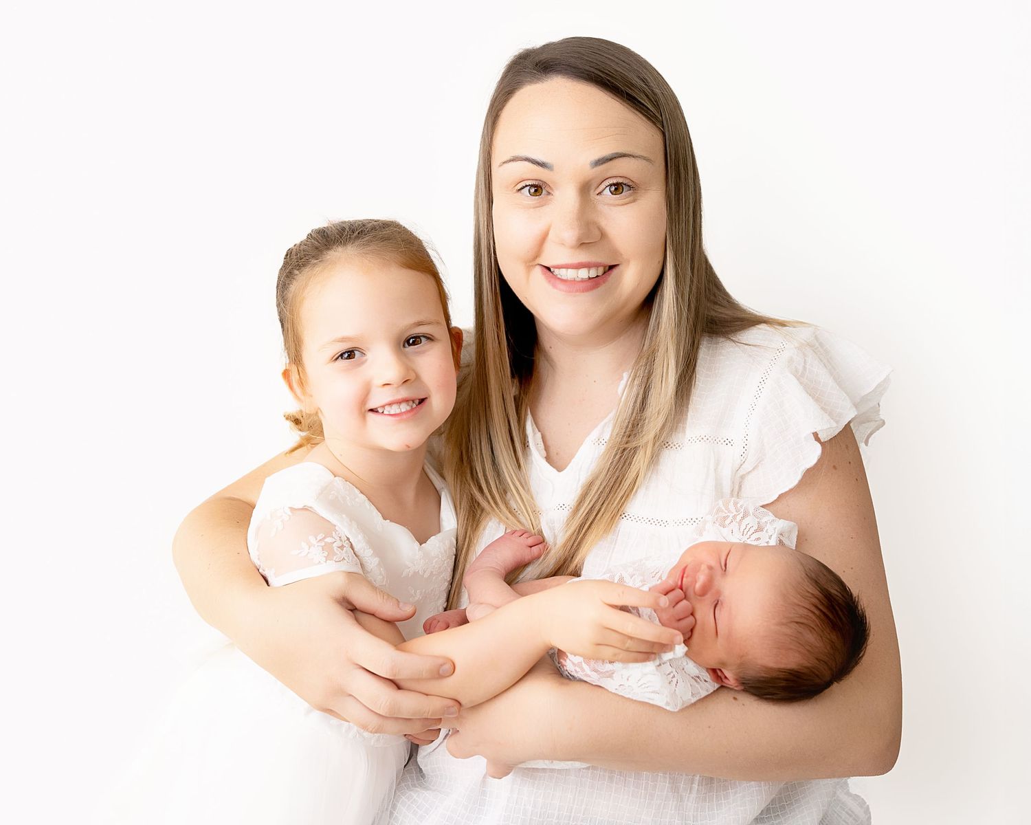 A mother in white holds a newborn baby while embracing a young child, all against a soft white background.