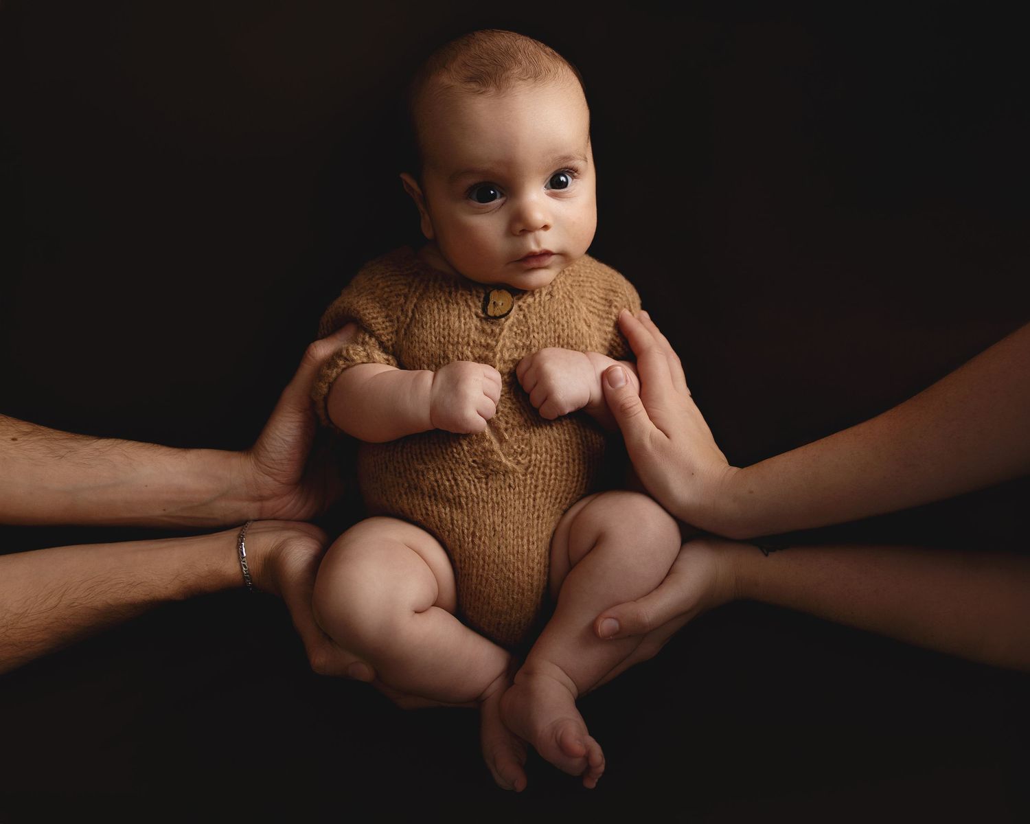 A baby wearing a brown knit romper is held safely by supporting hands against a dark backdrop.