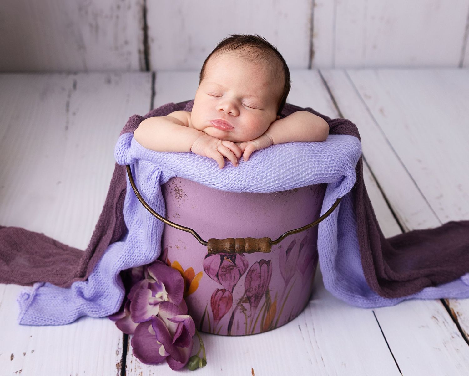 A sleeping newborn baby rests on a purple cushion inside a decorative bucket with floral designs and tulip accents.