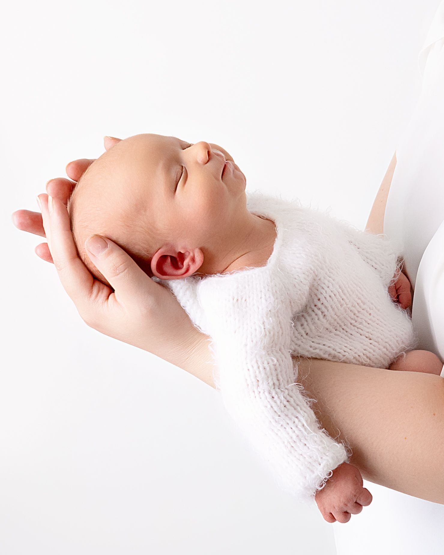A sleeping newborn infant in white clothing is gently held in supportive hands against a light background.