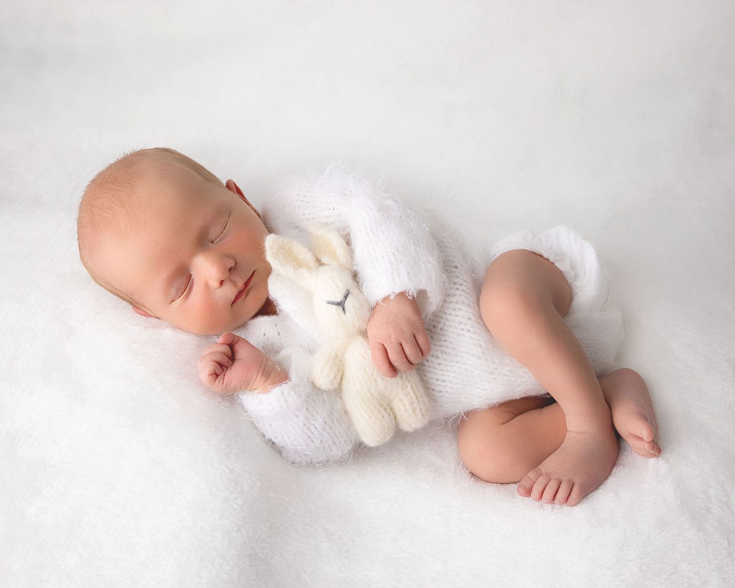 Sweet baby sleeping on white blanket while holding small cream-colored teddy bear plush toy.
