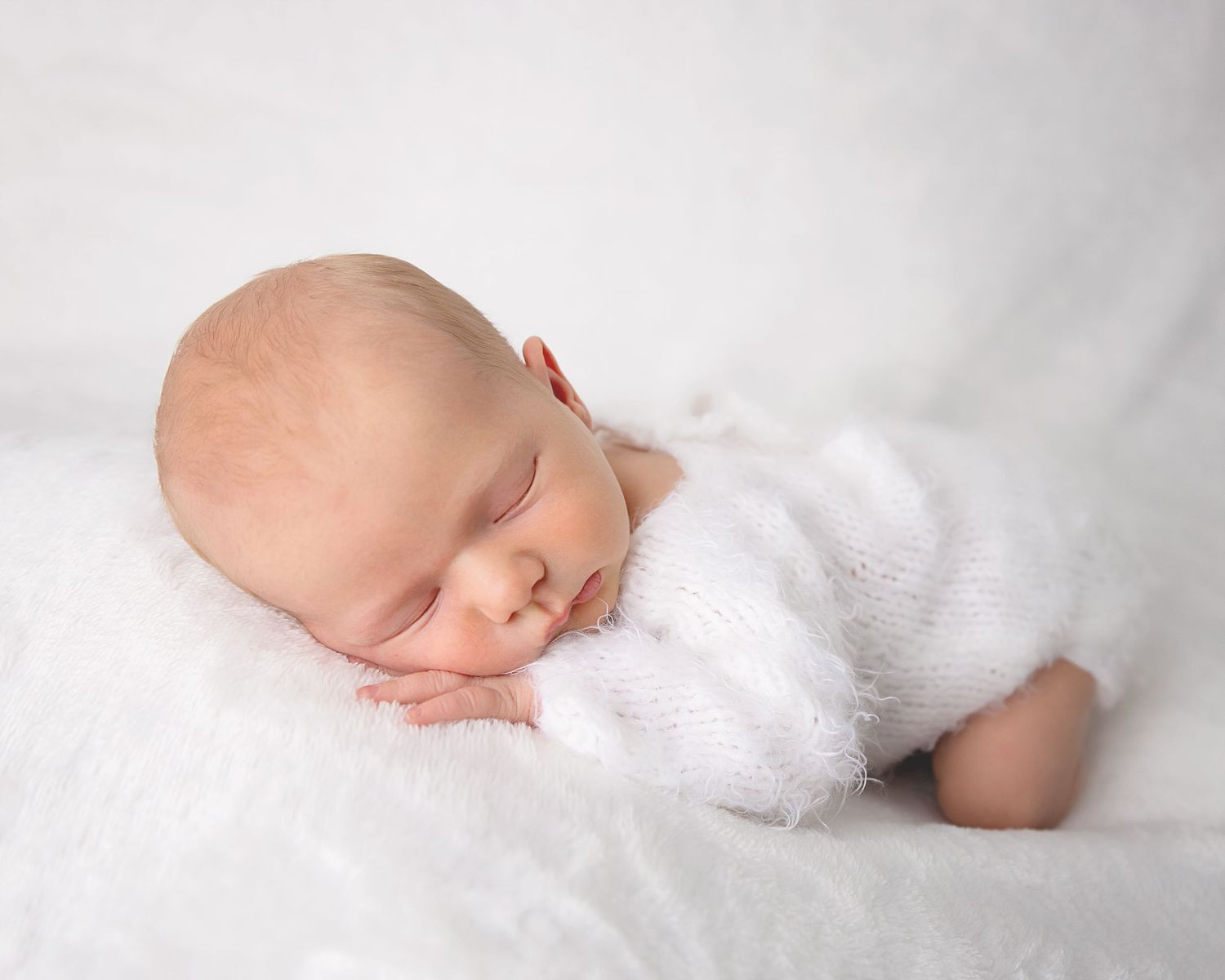 Peaceful newborn baby sleeping on soft white blanket wearing cozy white knit sweater.