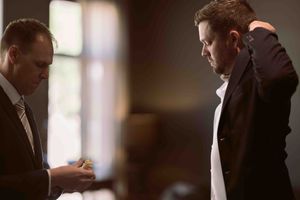 A man about to be married puts on jacket while wedding attendant snacks on a cake