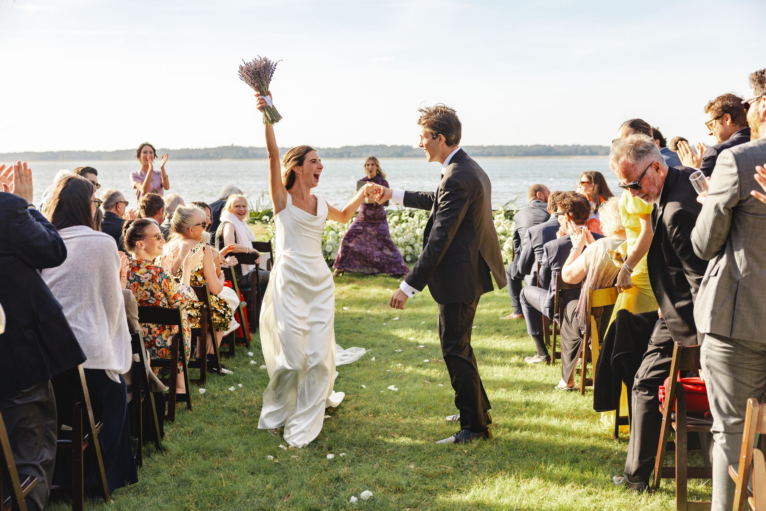 Wedding guests gather outdoors on a grassy lawn by the water during a sunny celebration.