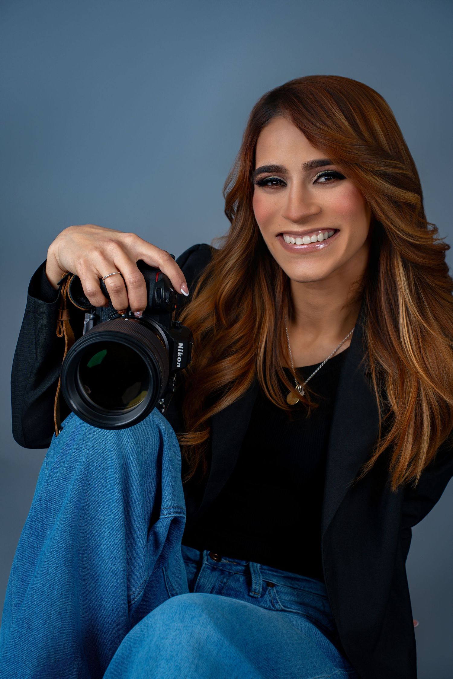 Professional photographer in black top and denim holding camera against blue backdrop.