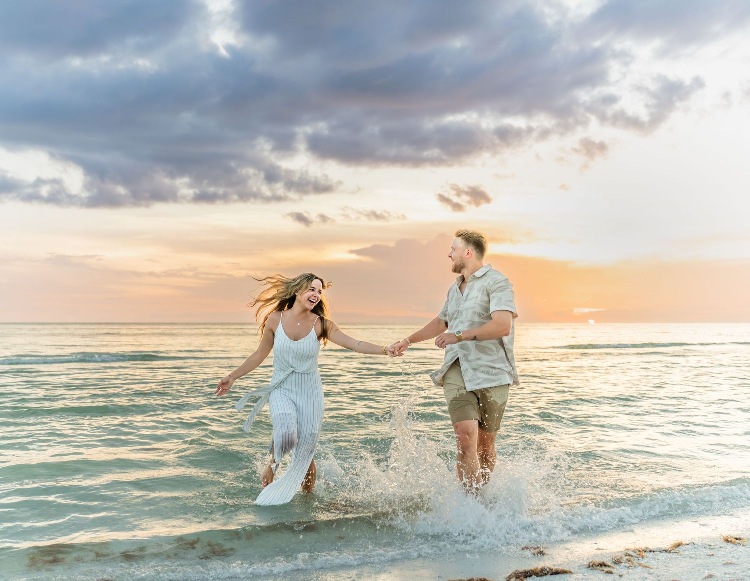 A playful couple splashing in ocean waves during sunset with dramatic sky.