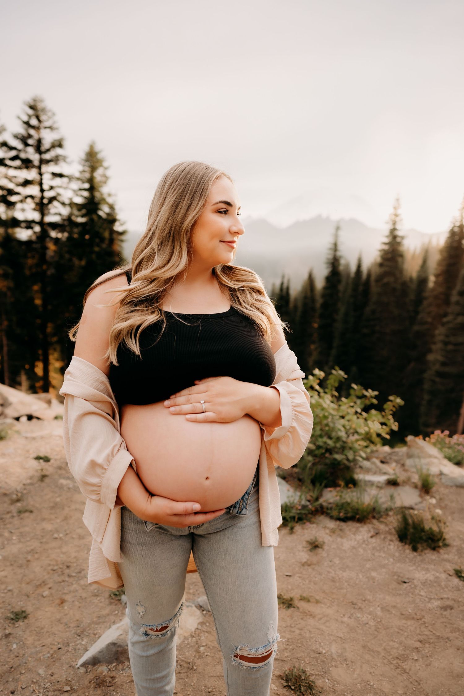 Tami smiles with her hands cradling her pregnant belly in maternity dress with a warm, summer sunset