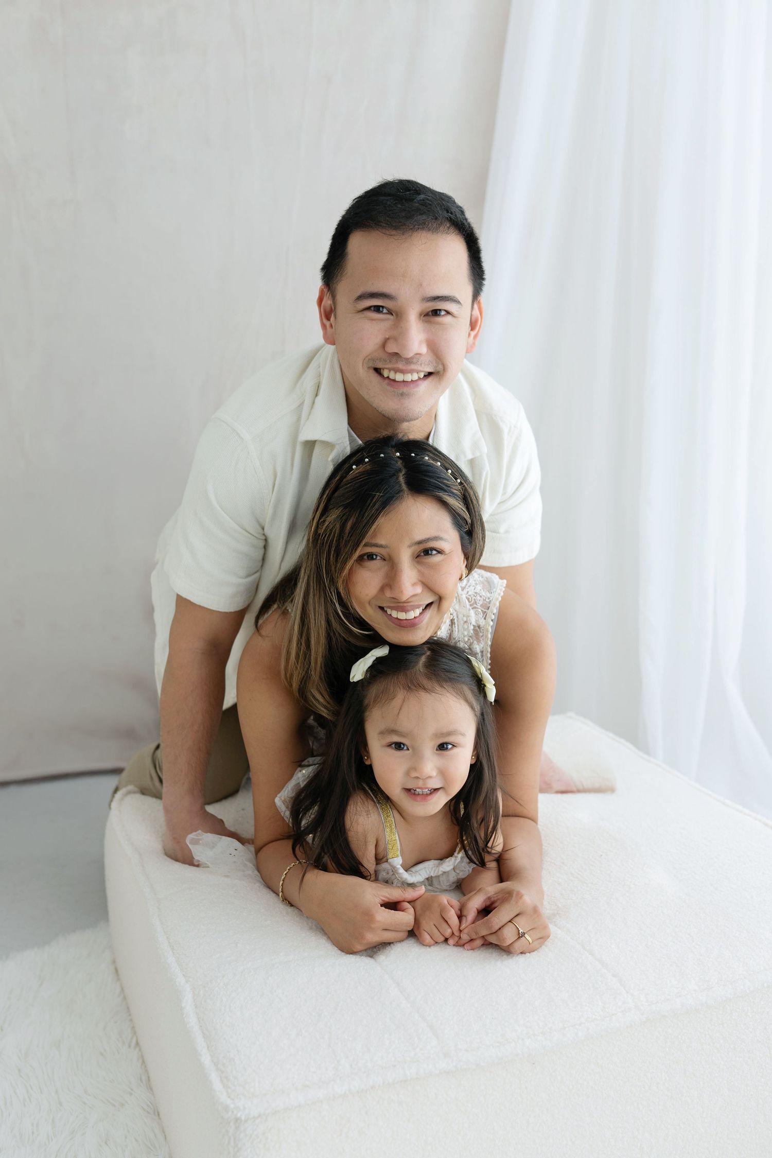 Happy Asian family of three posing together, parents and young daughter smiling in a bright white studio setting.