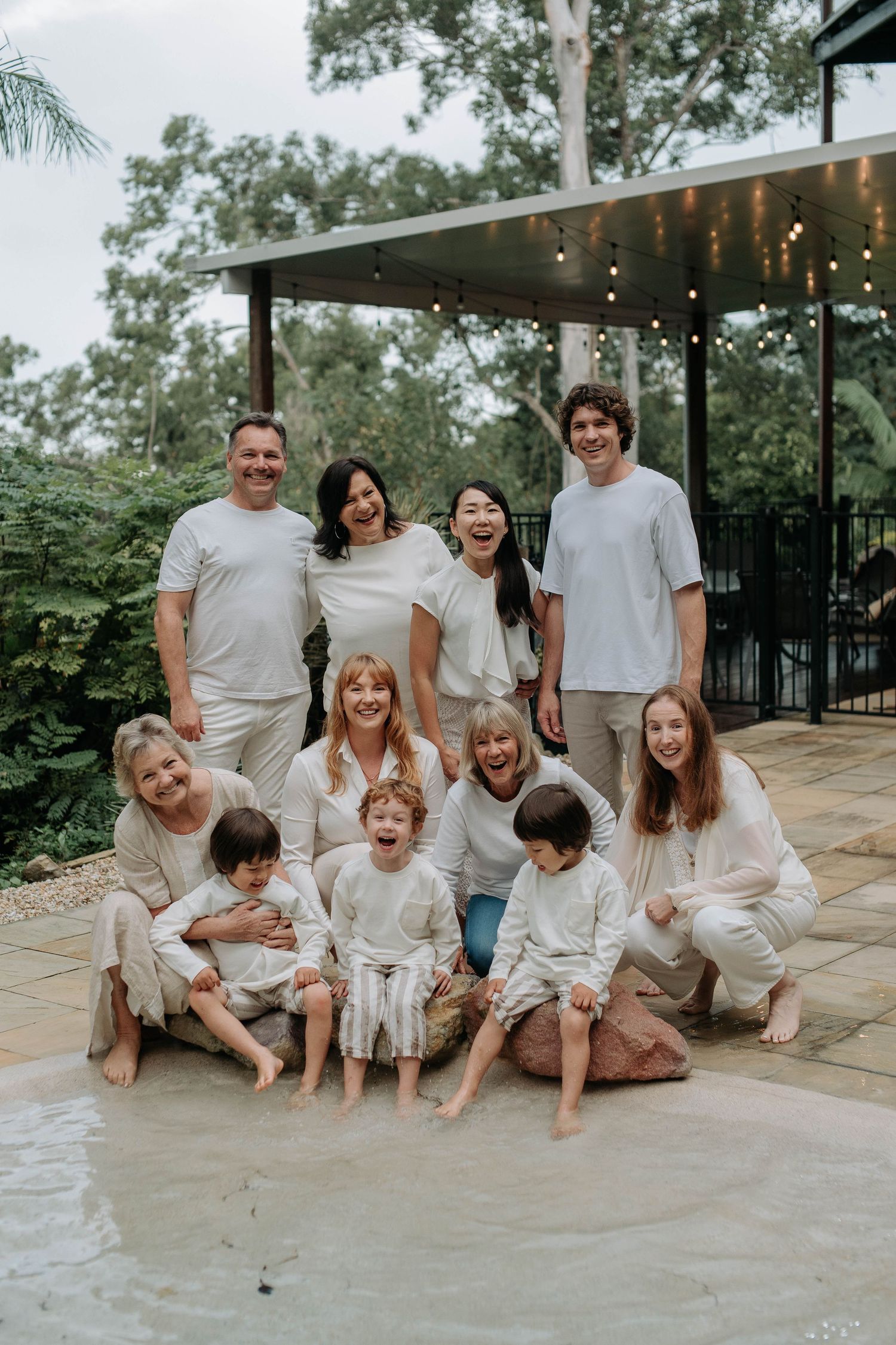 Large multigenerational family in matching white outfits posing and laughing together by a pool in a lush garden.
