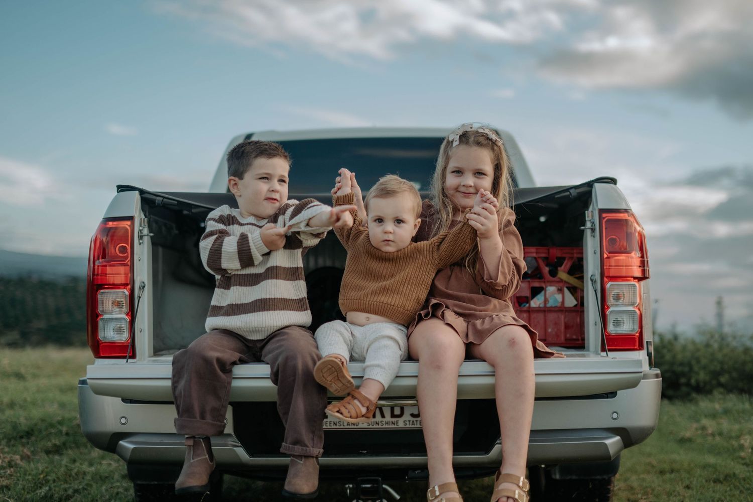 Three children sitting on a pickup truck tailgate outdoors, smiling and eating snacks against a cloudy sky.
