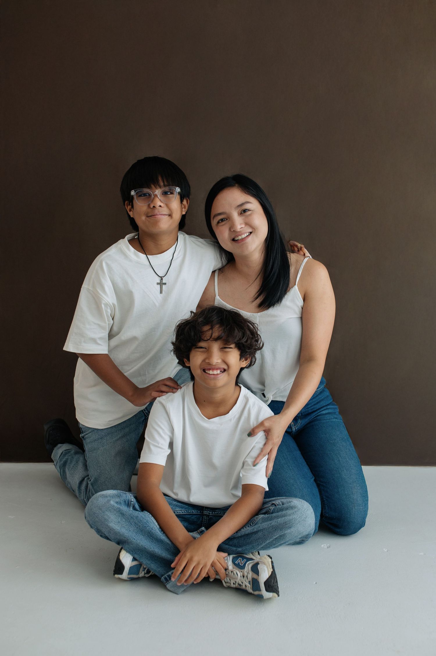 A woman and two boys in matching white shirts and jeans pose together for a family portrait against a brown background.