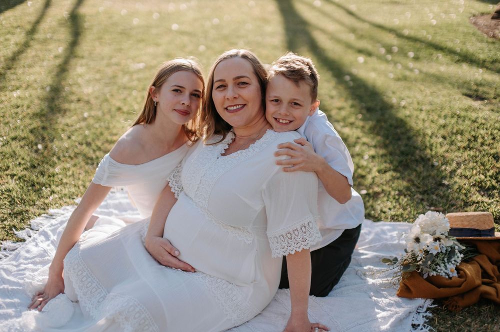 Pregnant woman in white dress sits on blanket outdoors with two children smiling during maternity photo session.