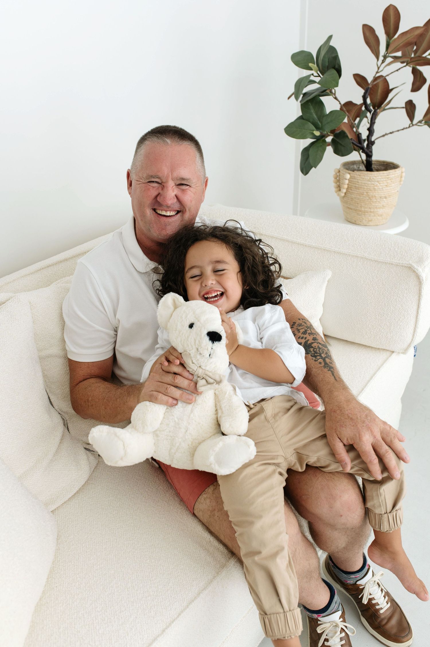 A man and young child laugh together on a white sofa, child holding a white teddy bear in a bright room.