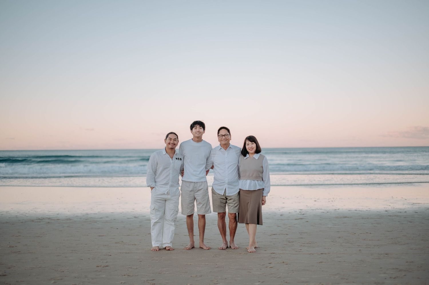 Family of four posing together on a sandy beach at sunset with calm ocean waves in the background.