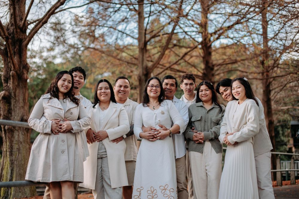 A joyful family group poses outdoors among autumn trees, dressed in elegant neutral and white outfits for a portrait.