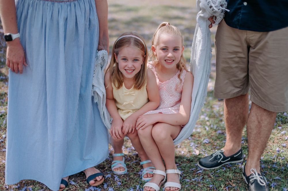 Two young girls smiling and posing together outdoors at a family gathering, wearing pastel dresses and white sandals.
