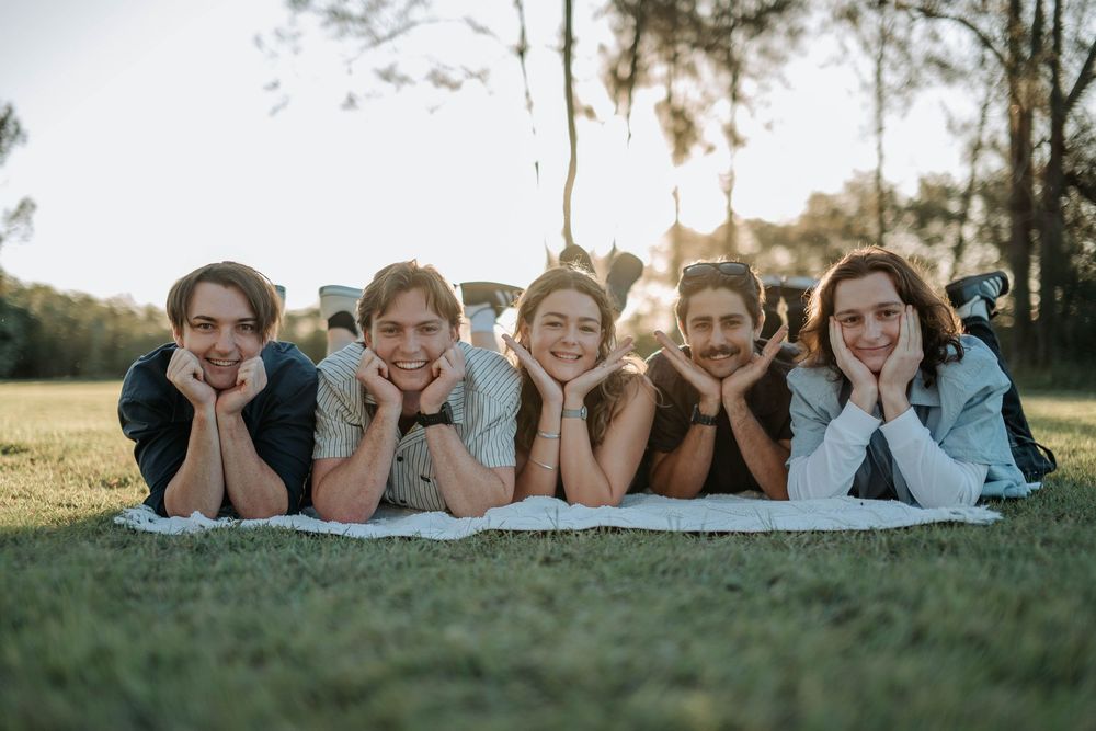 Five young friends lying on a blanket in a park, smiling with hands on chins during golden hour.