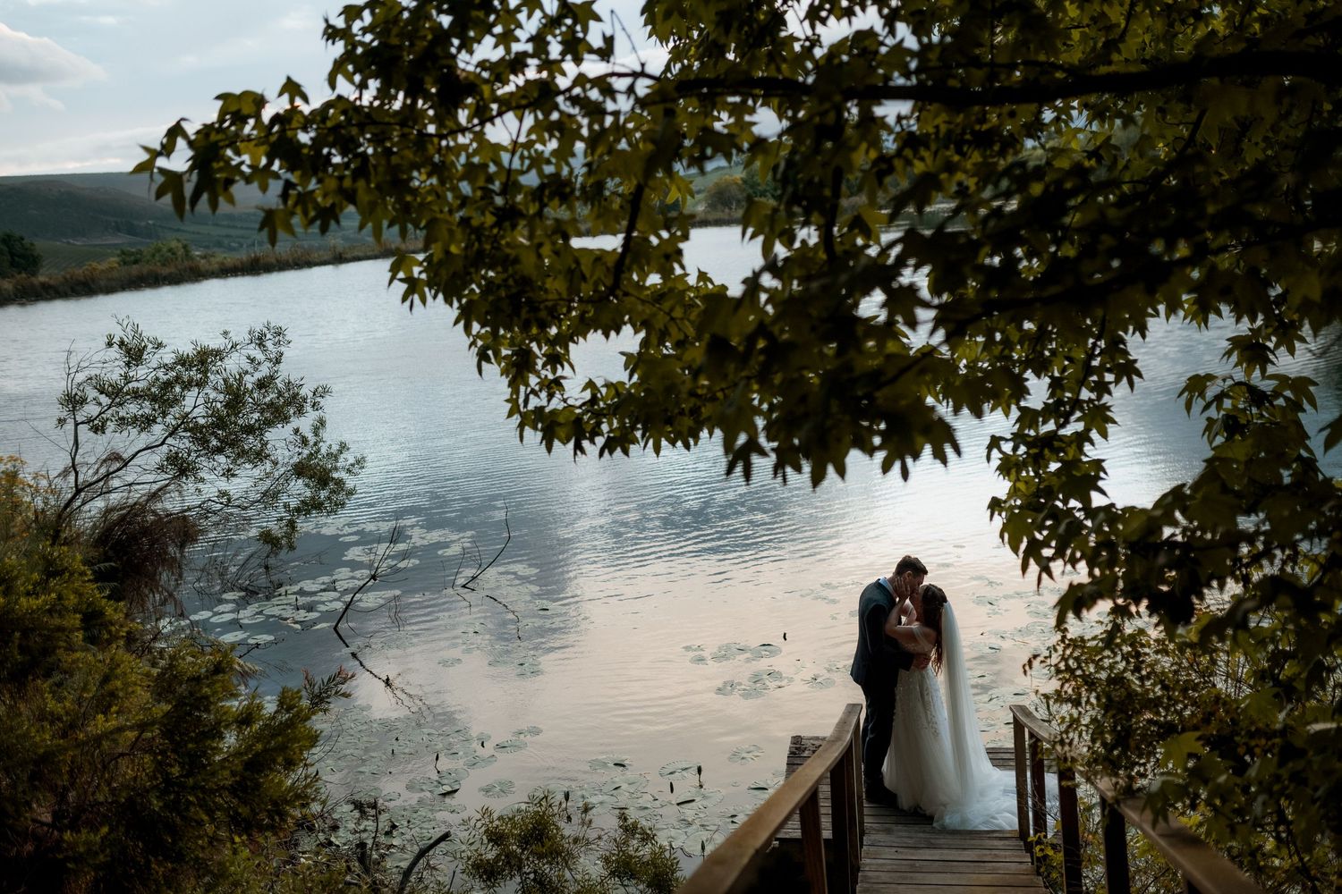Two silhouettes share a romantic moment on a wooden dock overlooking a serene lake at sunset framed by tree branches.