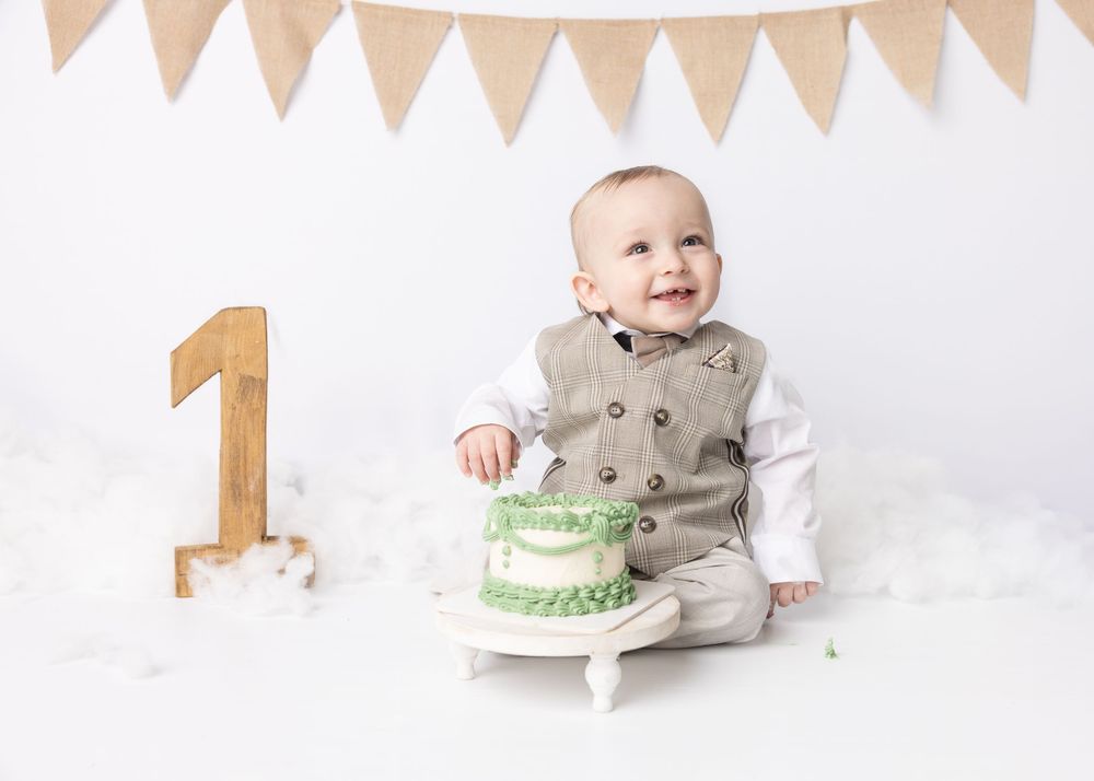 A small child in gray outfit sits on white backdrop with gold number one decoration and green birthday cake for cake smash photo.