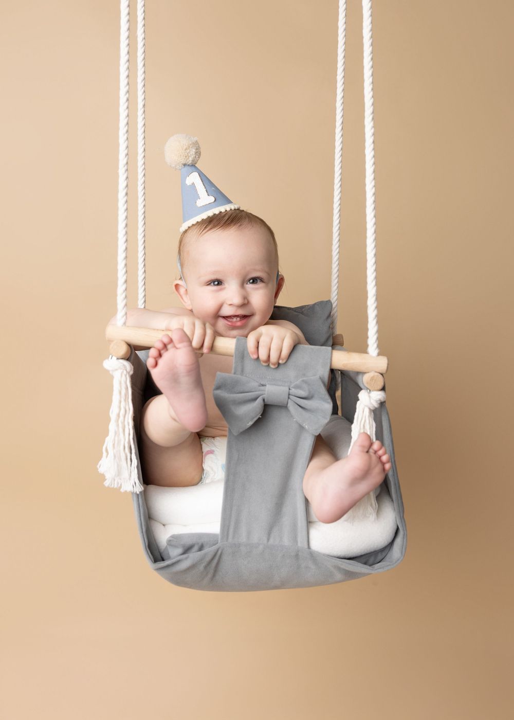 Baby wearing blue party hat sits in gray fabric swing with tassels against neutral background, smiling happily.