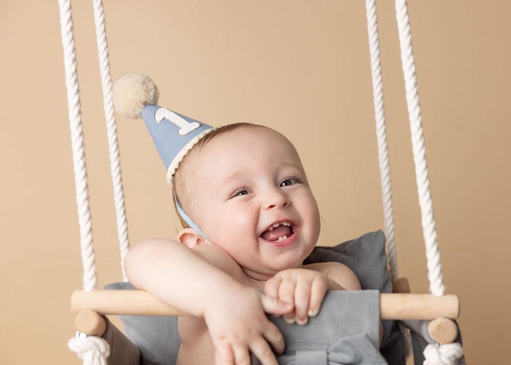 Happy baby wearing blue birthday hat sits in wooden swing during first birthday photoshoot.