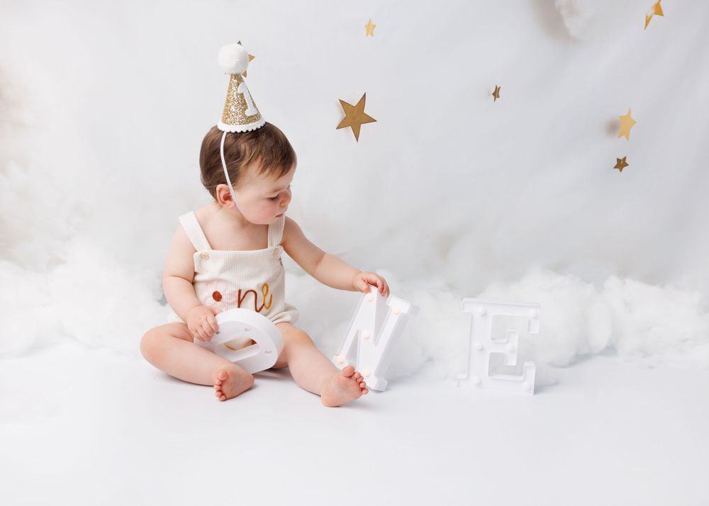 Baby in cream outfit plays with blocks on white cloudy backdrop with gold stars.