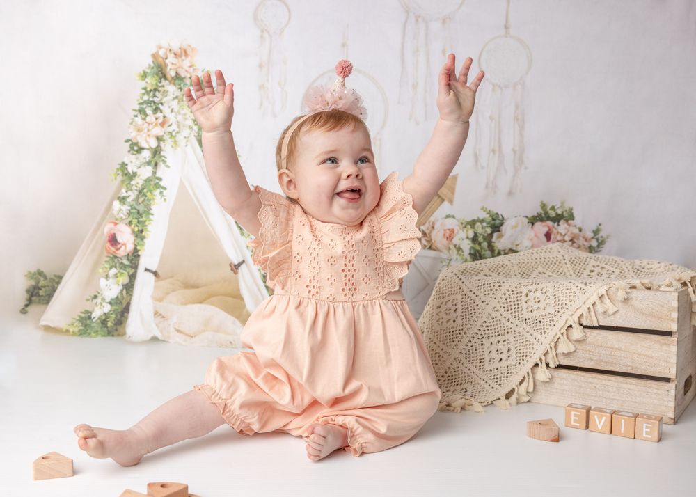 A joyful infant in a peach-colored dress smiles with raised arms against a light backdrop with floral accents.