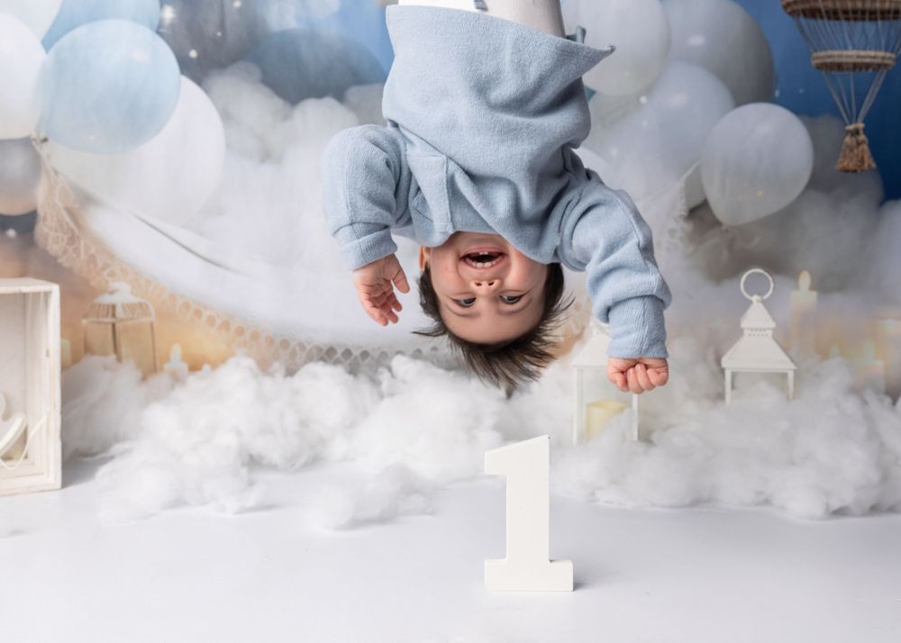 Creative upside-down photo of baby in blue hoodie against cloudy backdrop with number one decoration.