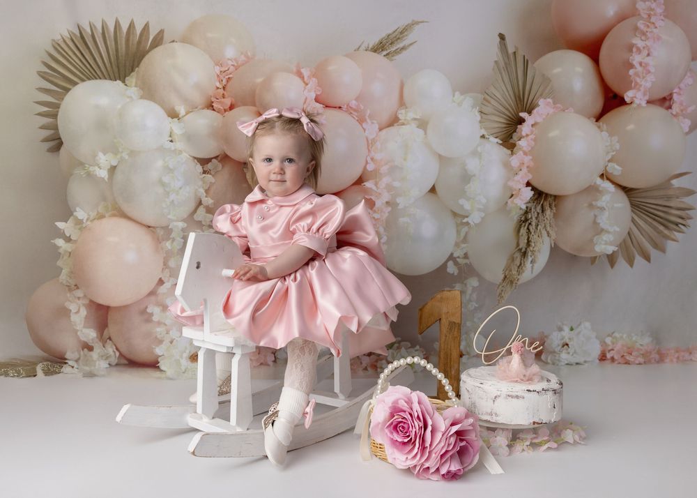 Elegant birthday setup with pink dress, rocking horse, roses and balloon garland in soft peach and cream colors.