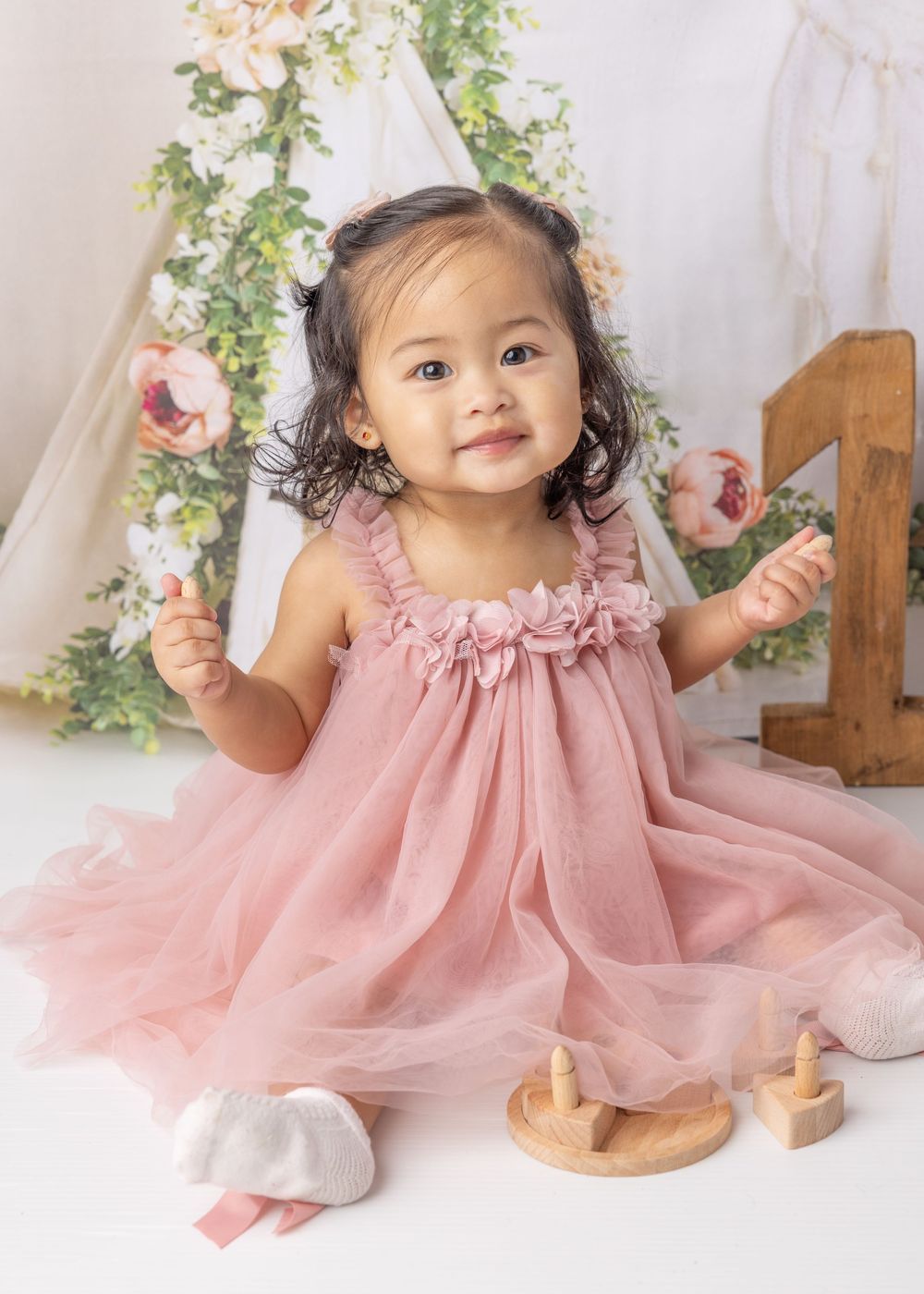 Sweet toddler wearing pink ruffled tulle dress poses with wooden toys amid floral decorations for a portrait session.