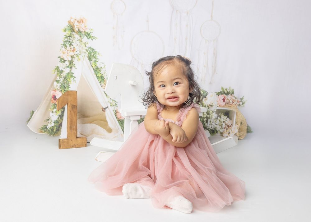 A baby wearing a pink tulle dress sits on a white backdrop with a floral decorated teepee and basket in the background.