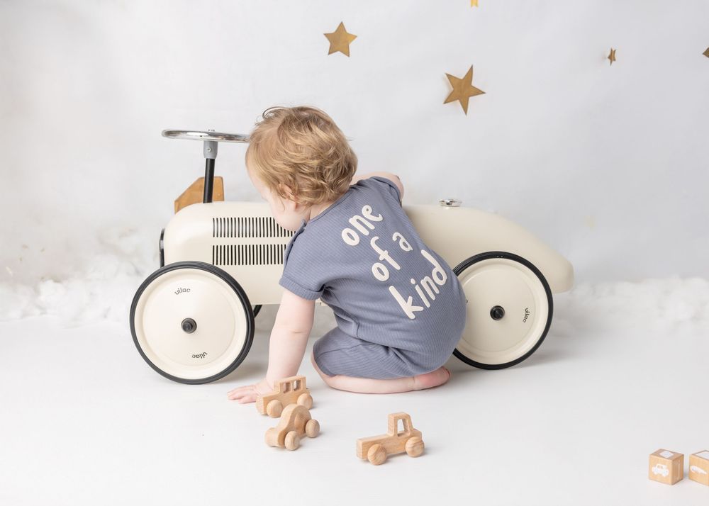 Small child in grey romper plays with wooden toys near vintage-style ride-on car against white backdrop with gold stars.