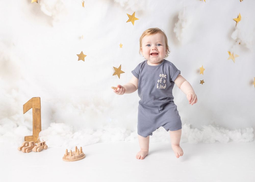 Baby wearing grey romper sits on white backdrop with gold star decorations for first birthday milestone photo.