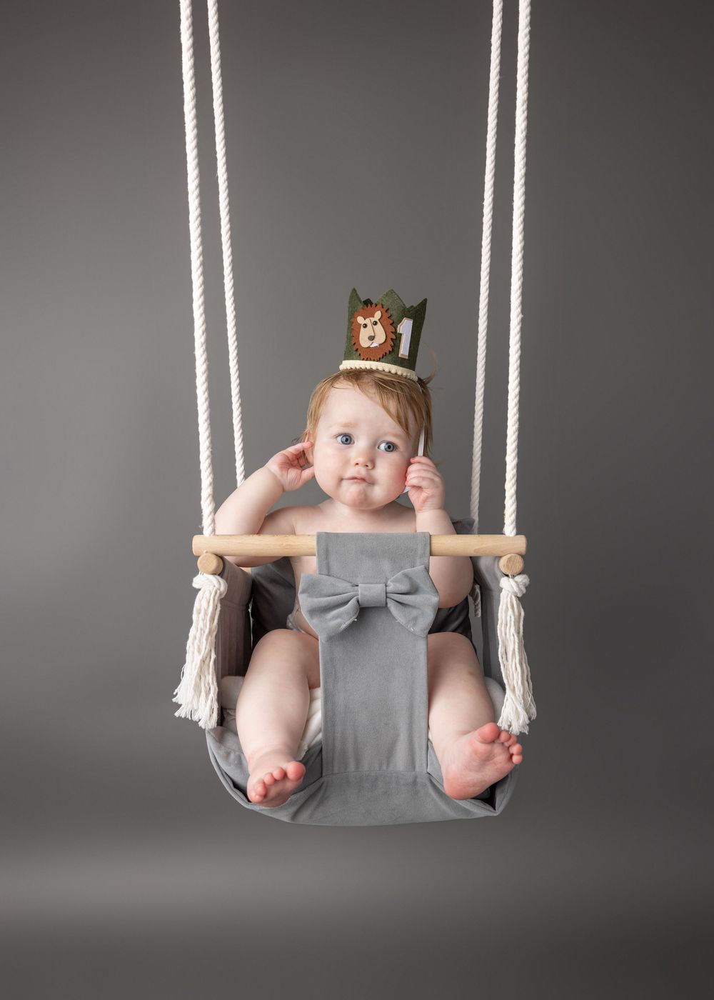 Adorable baby wearing a crown and gray romper swing gleefully in a rope swing against a gray studio backdrop.