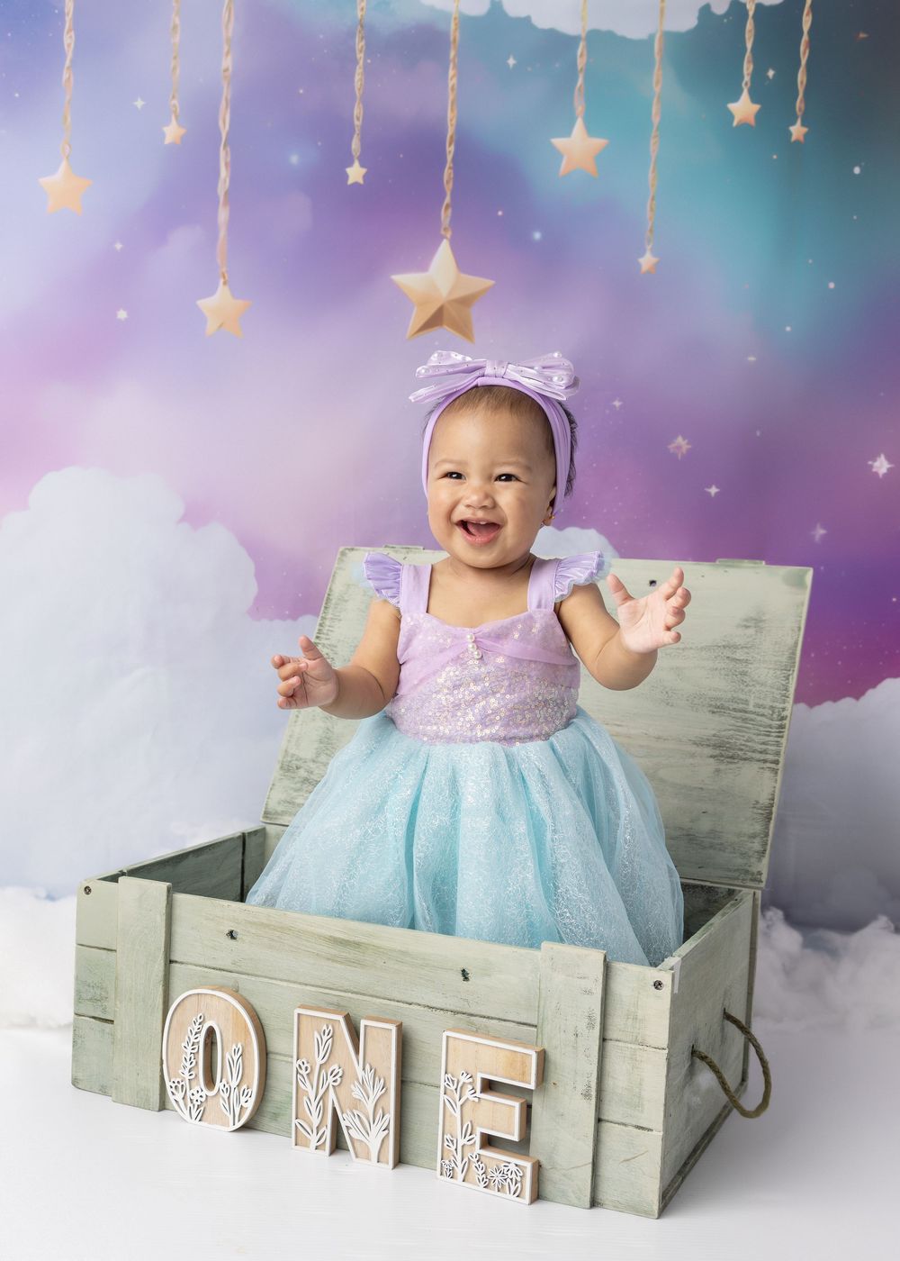 A festive first birthday photo setup with a mint and lavender backdrop featuring hanging stars and a rustic wooden crate.