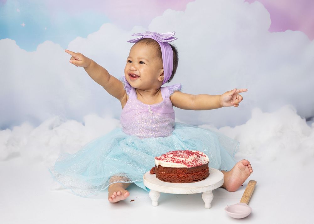 Baby wearing purple bow and tulle dress poses with chocolate birthday cake on fluffy cloud backdrop.