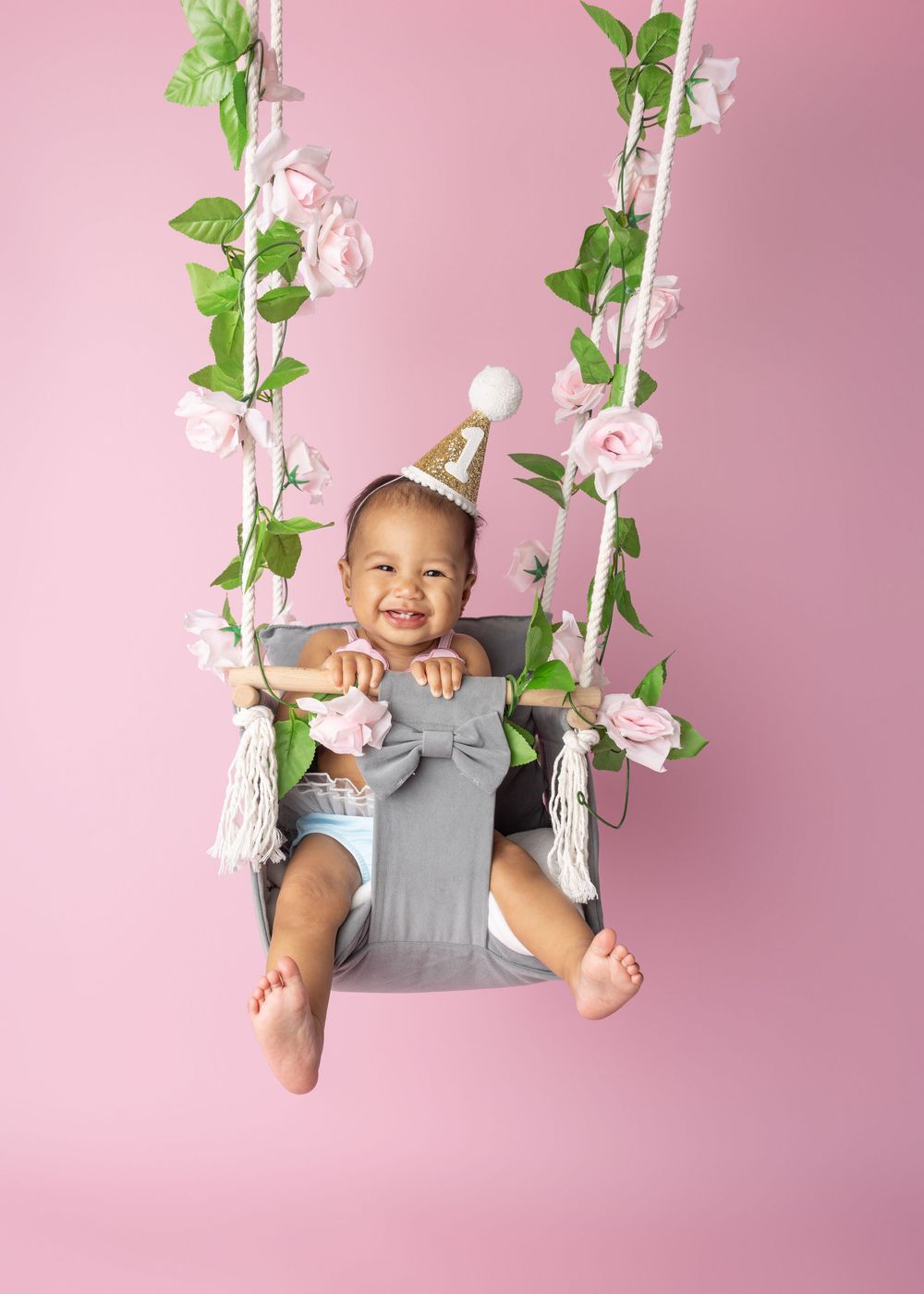 Festive scene with baby sitting in a gray swing decorated with delicate pink roses and green vines against pink backdrop.