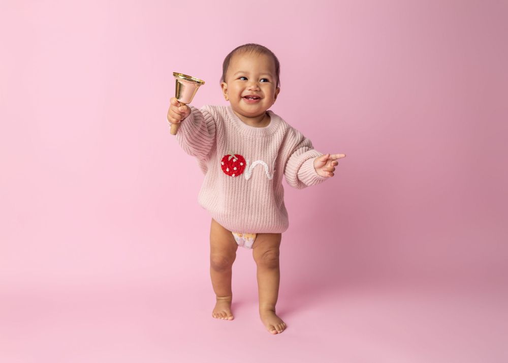 Baby wearing pink knit sweater stands and smiles against pink background.