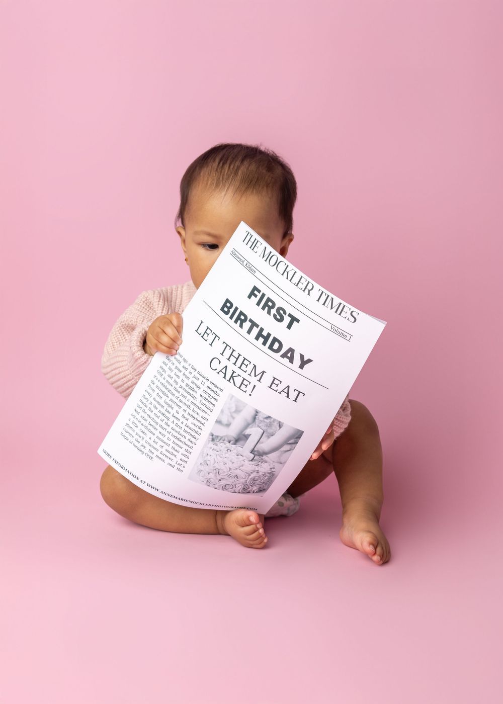 Baby holding a newspaper with 'First Birthday' headline sits against pink backdrop.